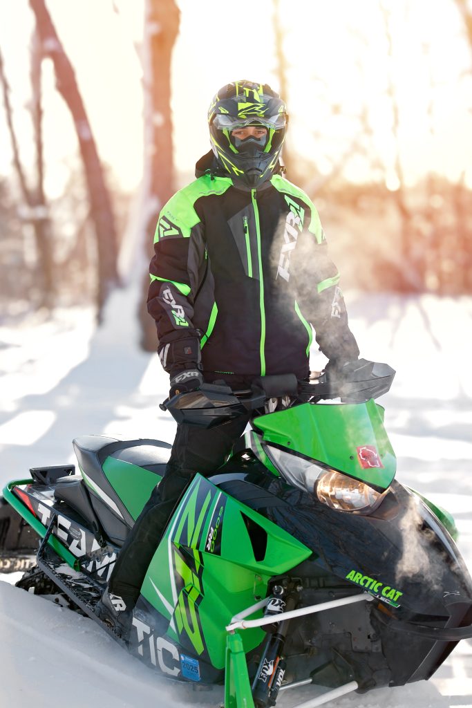 High school senior boy winter portrait by Teri Lynn Photography in Brodhead, WI. Young man in a neon green and black snowmobile suit standing on a green Arctic Cat snowmobile in a snowy forest, captured with soft natural light. Senior photography highlighting winter sports and personal interests.