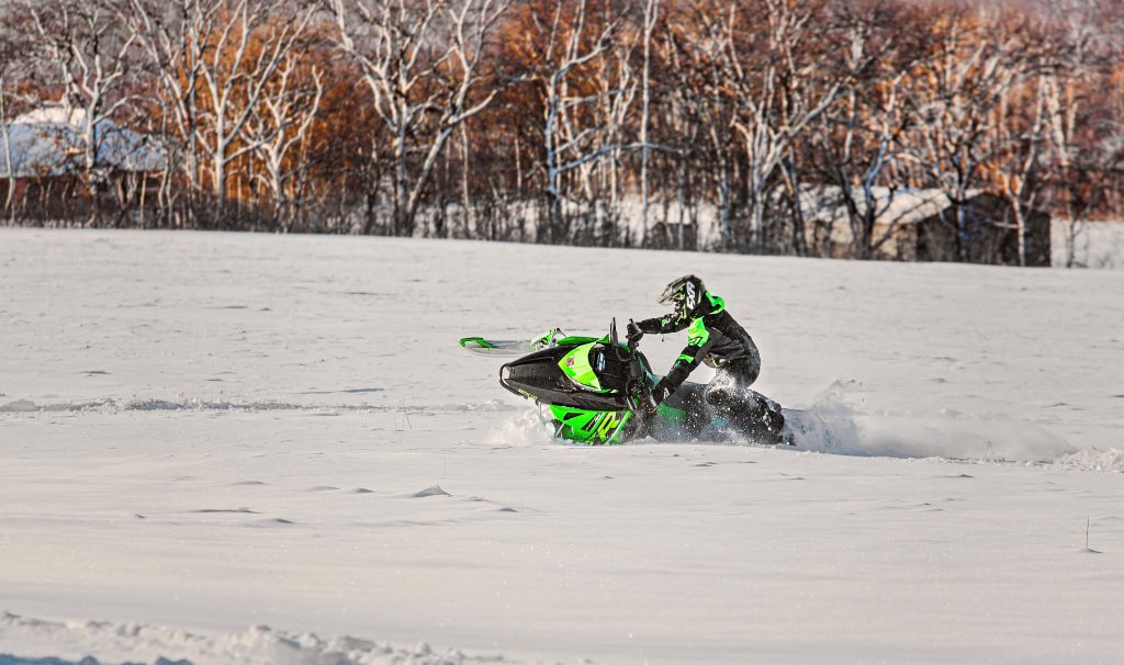 High school senior boy action shot by Teri Lynn Photography in Brodhead, WI. Young man in a neon green and black snowmobile suit riding a green Arctic Cat snowmobile in a snowy field, with a backdrop of trees. Dynamic senior photography showcasing winter sports and outdoor activities.