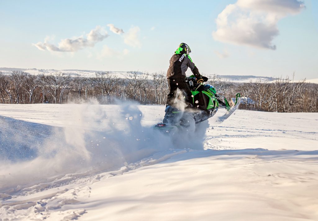 High school senior boy snowmobiling by Teri Lynn Photography in Brodhead, WI. Young man in a neon green and black snowmobile suit performing a maneuver on a green Arctic Cat snowmobile, with snow flying behind. Action-packed senior photography capturing winter sports and adventures.