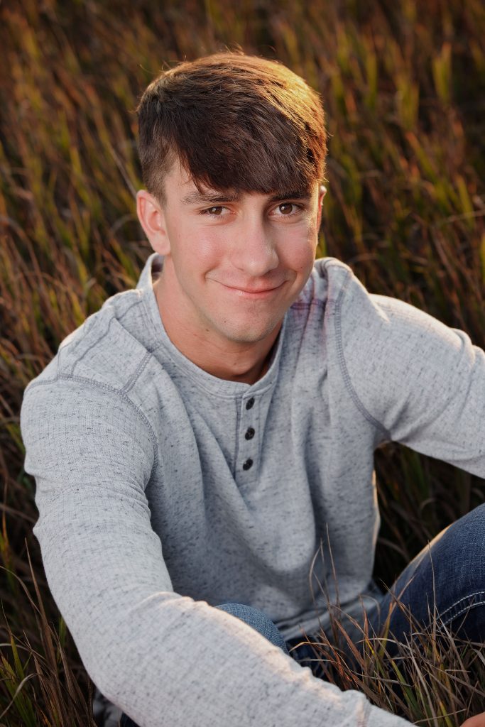 High school senior boy outdoor portrait by Teri Lynn Photography in Brodhead, WI. Smiling young man in a gray henley shirt sitting in tall grass during golden hour. Professional senior photography specializing in natural settings