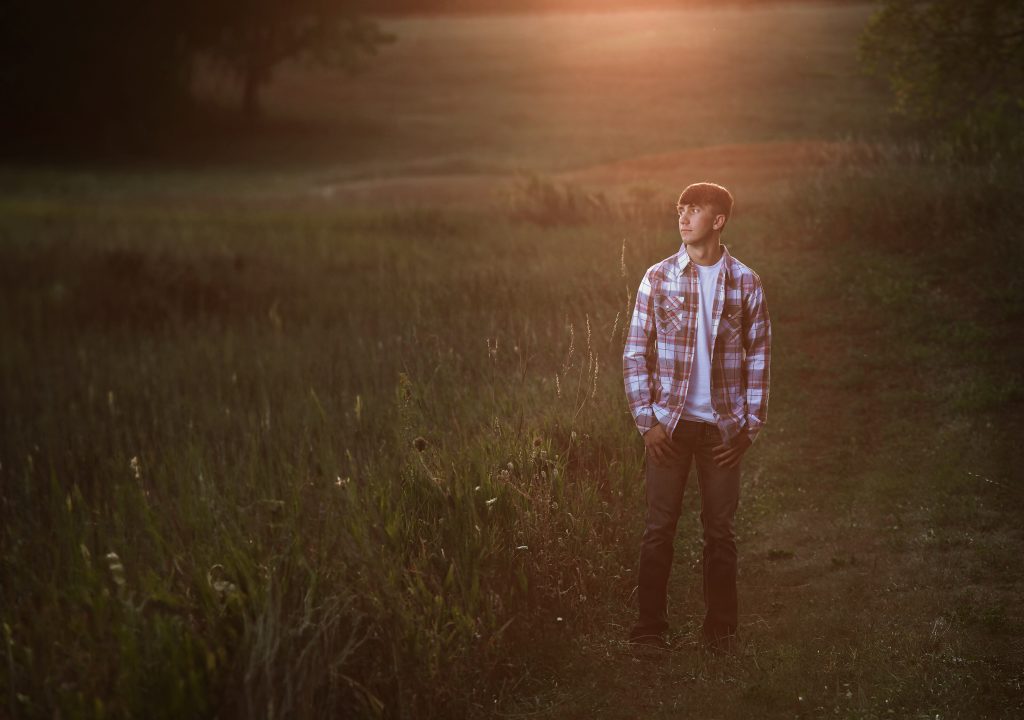 High school senior boy outdoor portrait by Teri Lynn Photography in Brodhead, WI. Young man in a plaid shirt standing in a field of tall grass at sunset, looking off into the distance. Professional senior photography capturing natural and relaxed moments.