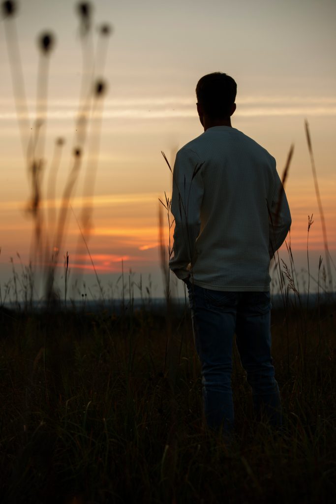 High school senior boy sunset silhouette by Teri Lynn Photography in Brodhead, WI. Young man in a light sweater standing in a field, backlit by a colorful sunset. Creative senior portraits showcasing dramatic and serene outdoor settings