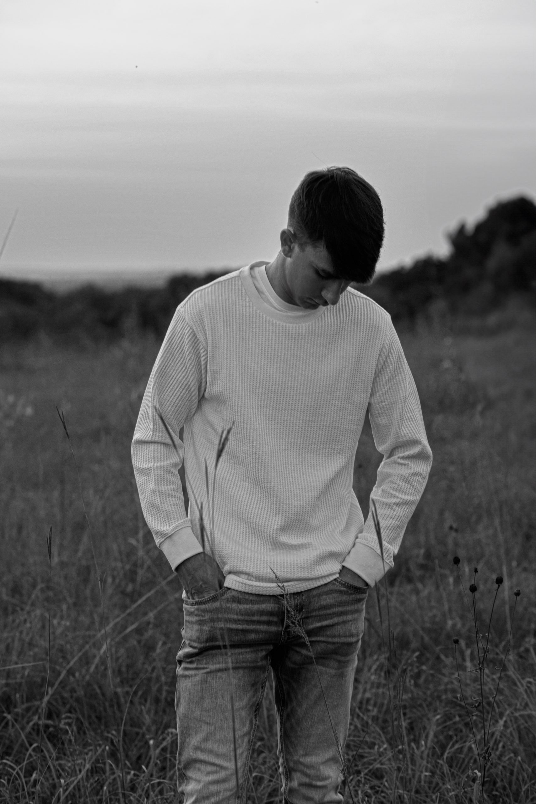 Black and white high school senior portrait by Teri Lynn Photography in Brodhead, WI. Young man in a light sweater looking down, standing in a field. Artistic senior photography with a focus on natural emotions and settings