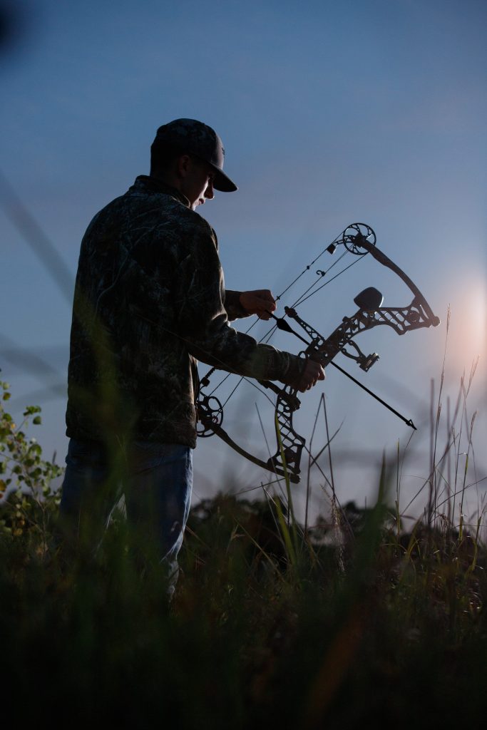 High school senior boy outdoor portrait by Teri Lynn Photography in Brodhead, WI. Silhouette of a young man in camouflage clothing adjusting his compound bow at dusk. Unique senior photography showcasing personal interests and hobbies.