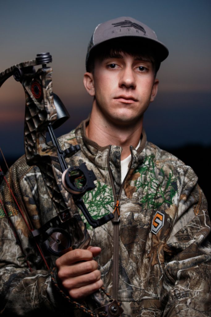 High school senior boy close-up portrait by Teri Lynn Photography in Brodhead, WI. Young man in camouflage jacket holding a compound bow, looking directly at the camera with a serious expression. Professional senior photography that captures personality and interests