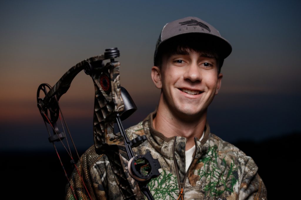 High school senior boy close-up portrait by Teri Lynn Photography in Brodhead, WI. Young man in camouflage jacket holding a compound bow, smiling warmly at the camera during an evening photo session. Senior photography emphasizing natural expressions and personal interests