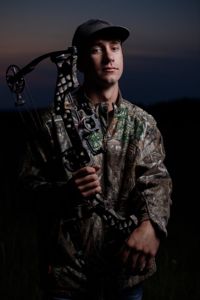 High school senior boy portrait by Teri Lynn Photography in Brodhead, WI. Young man in camouflage jacket holding a compound bow, standing confidently during an evening photo session. Professional senior photography capturing hobbies and unique personalities