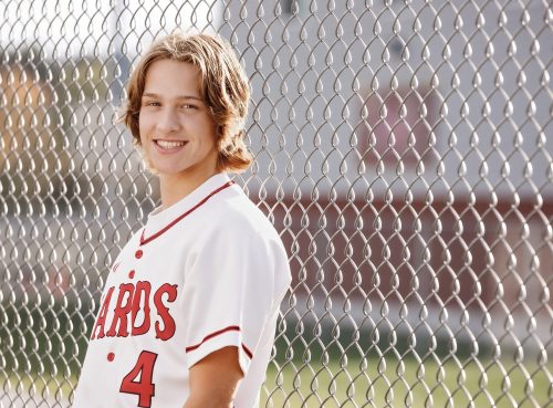 High school senior boy leaning against a fence on the baseball field wearing his school jersey at Brodhead High School in Wisconsin