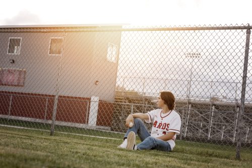 High school senior boy leaning against a fence on the baseball field wearing his school jersey at Brodhead High School in Wisconsin