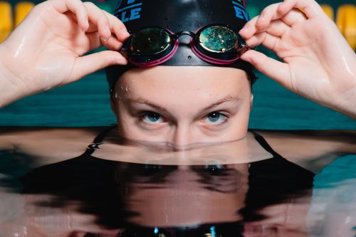 High school senior swimmer portrait by Teri Lynn Photography in Janeville, WI. Parker High School. Young woman wearing a swimming cap and goggles, partially submerged in a pool, looking directly at the camera. Professional senior photography highlighting athleticism and dedication